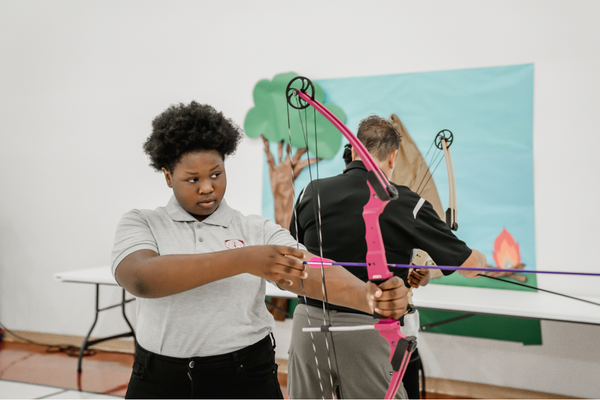 PACE Academy scholar sets up a purple arrow on a pink bow while a teacher shows another student how to maneuver bow in the background.