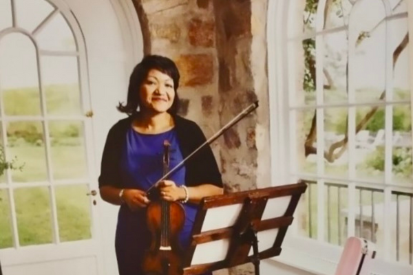 PACE Academy Strings teacher Angela Lott stands holding a violin and bow inside a stone building with large rounded windows.