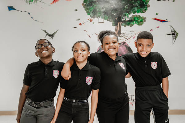 Four Pace Academy students in black uniform polo shirts smile in a row facing the camera with their arms around each other’s shoulders in front of a mural of a tree with objects that fly.