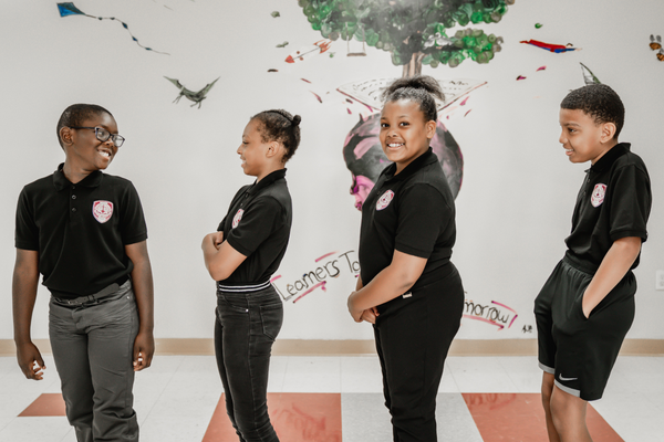 Four PACE Academy students stand in line in a hallway with a mural on the wall asserting their school motto, “Learners Today, Leaders Tomorrow”. One of the students smiles at the camera, two students look in front of them in line and student at the front looks behind him, smiling.