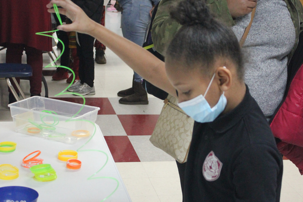 PACE Academy student reads some instructions while stretching a green spiraling plastic cable at a table with adults standing in the background.