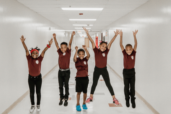 Five PACE Academy students in matching maroon shirts and black pants jump into the air in the school hallway with their arms outstretched over their heads.