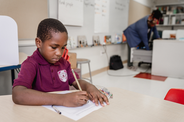 PACE Academy student sits at desk in classroom writing on a paper with a pencil. A teacher is just visible in the background.