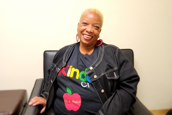 PACE Academy Family & Student Support Coordinator Ms. Ava Mullen sits in a chair in a black jacket and black shirt with an apple with her name on it.