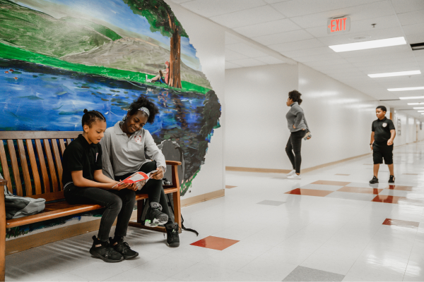Two PACE Academy students sit on a bench under a mural in a school hallway, one reading a book and the other student listening with a smile on their face.