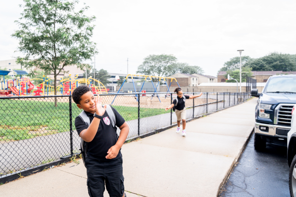 Two PACE Academy students wave goodbye on a sidewalk next to the large and colorful playground.