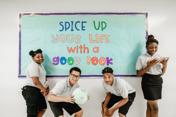 PACE Four Academy students stand in front of an turquoise bulletin board with the words “Spice Up Your Life with a Good Book” written on it in colored markers.