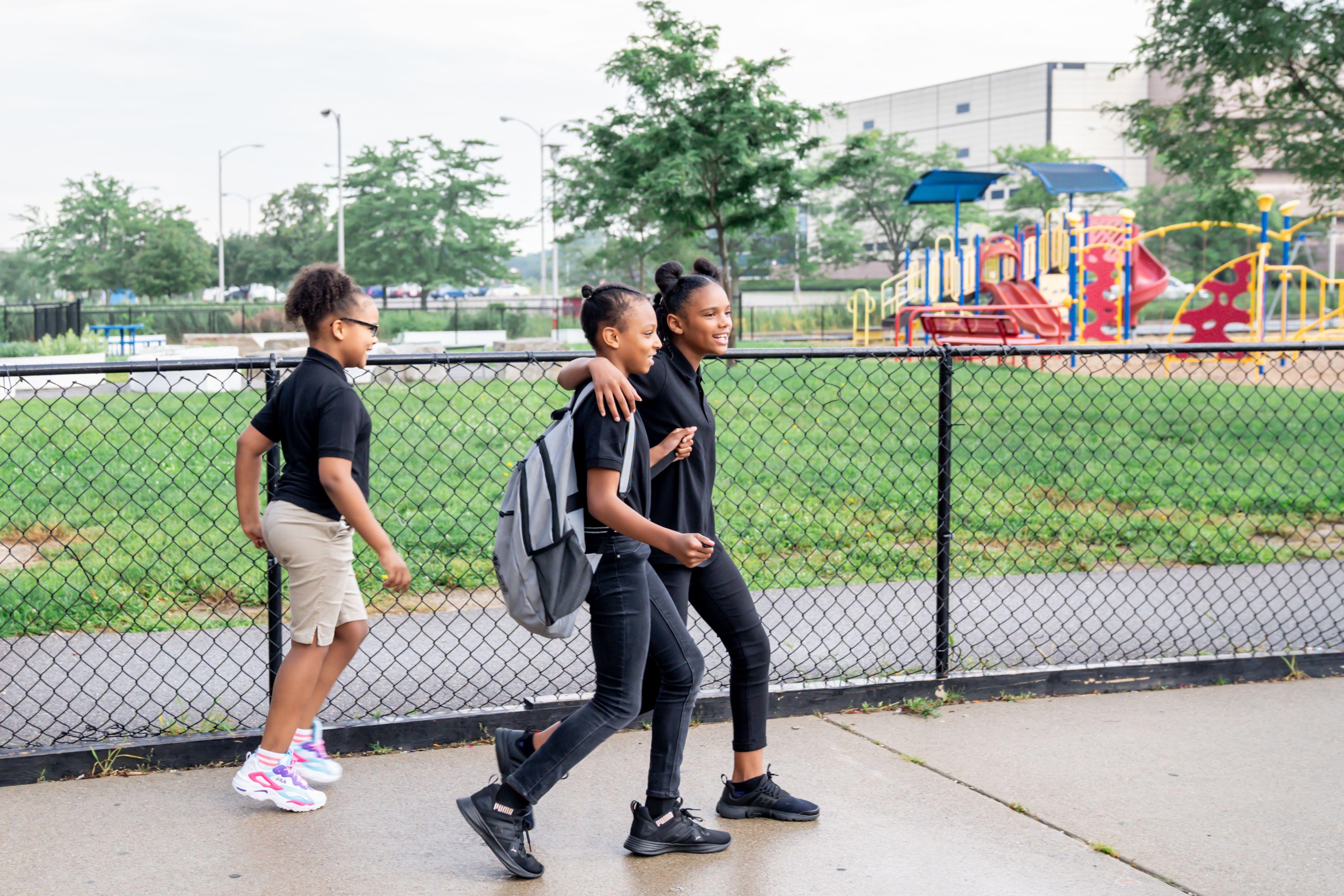 Outside PACE Academy, two students walk together smiling on a sidewalk near the colorful playground while another follows right behind.